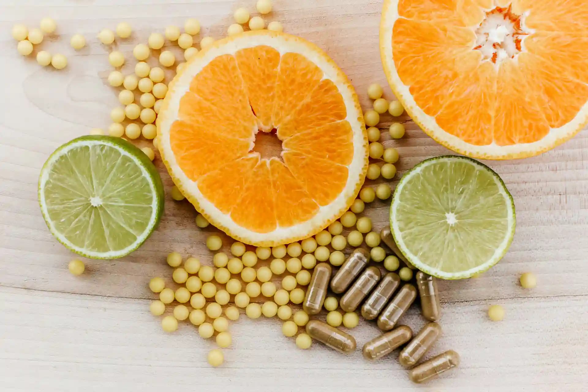 Ahşap bir zeminde portakal ve misket limonu dilimlerinin yanında duran sarı takviye taneleri ve bitkisel kapsüller